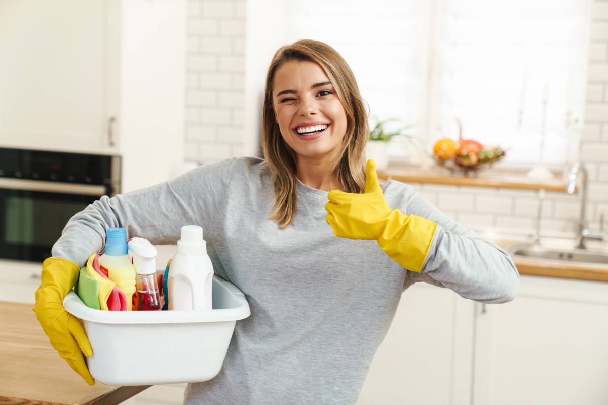 A woman in a gray sweatshirt and yellow rubber gloves holding a bin of cleaning supplies  grins and winks while giving a thumbs up.