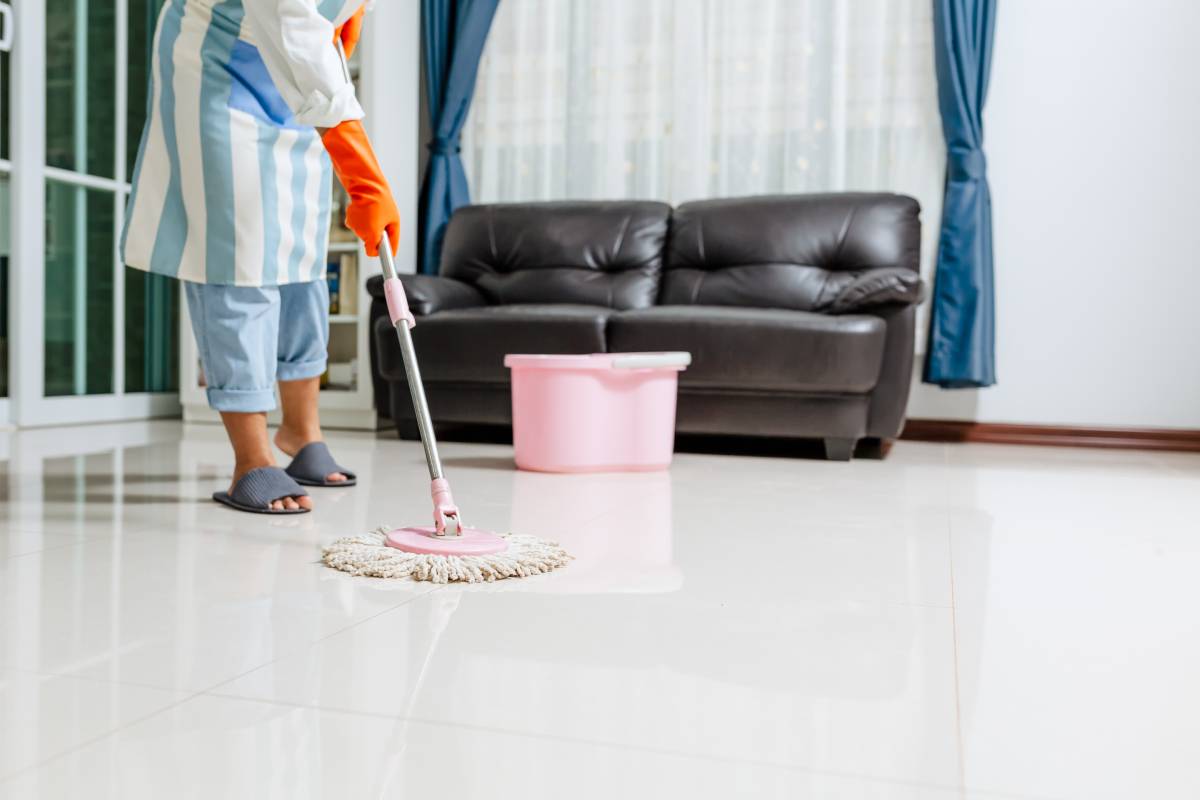 A woman in a striped apron and rubber gloves is seen from the torso down, using a spin mop and bucket to clean a white floor.